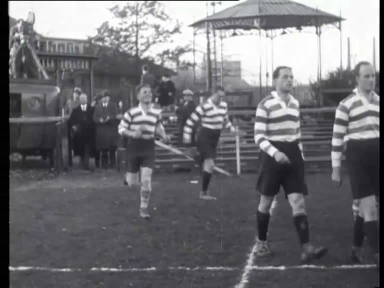Zwart-witfilmbeeld uit 1933 van rugbyspelers in shirts met horizontale banen die het veld betreden vóór AAC tegen AVRC in Amsterdam.