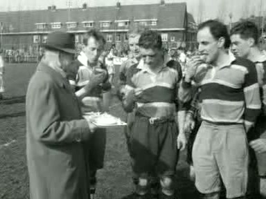Zwart-wit journaalbeeld van vijf AAC-rugbyspelers in shirts met brede banen die langs het veld iets drinken, Amsterdam april 1955.