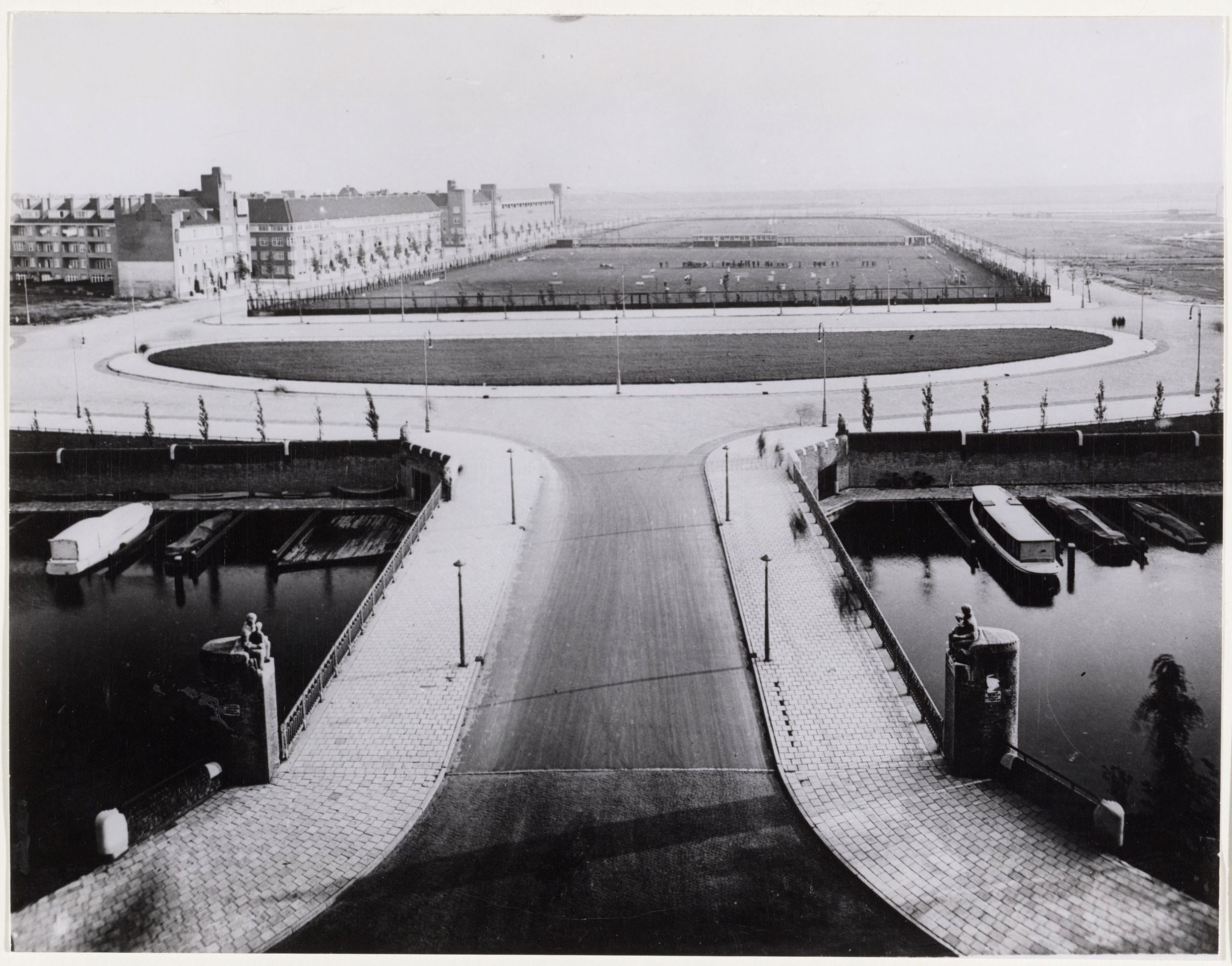 Zwart-witfoto over een brug richting het sportterrein Olympiaplein in mei 1932, met een atletiekbaan en middenveld achter de weg.