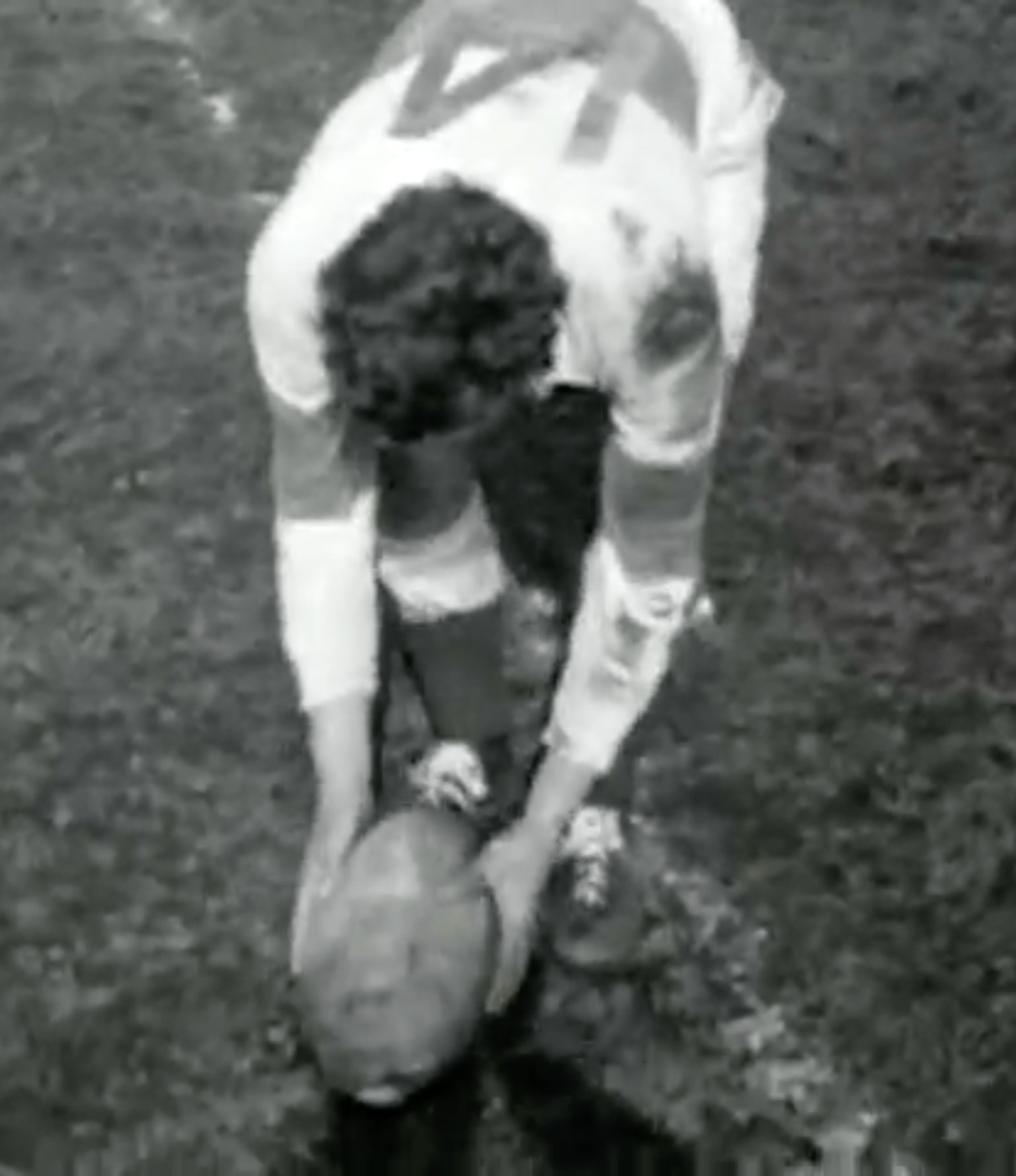 Zwart-wit close-up van een AAC-speler in een wit shirt met AAC op de rug, voorovergebogen terwijl hij een rugbybal van het gras pakt.