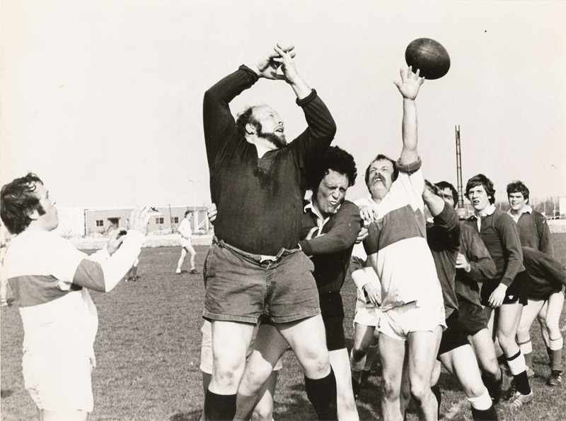 Zwart-witfoto van een rugby-line-out uit de jaren zeventig: een bebaarde AAC-speler springt naar de bal tegen tegenstanders in witgehoopte shirts, met het clubhuis en een lichtmast op de achtergrond.