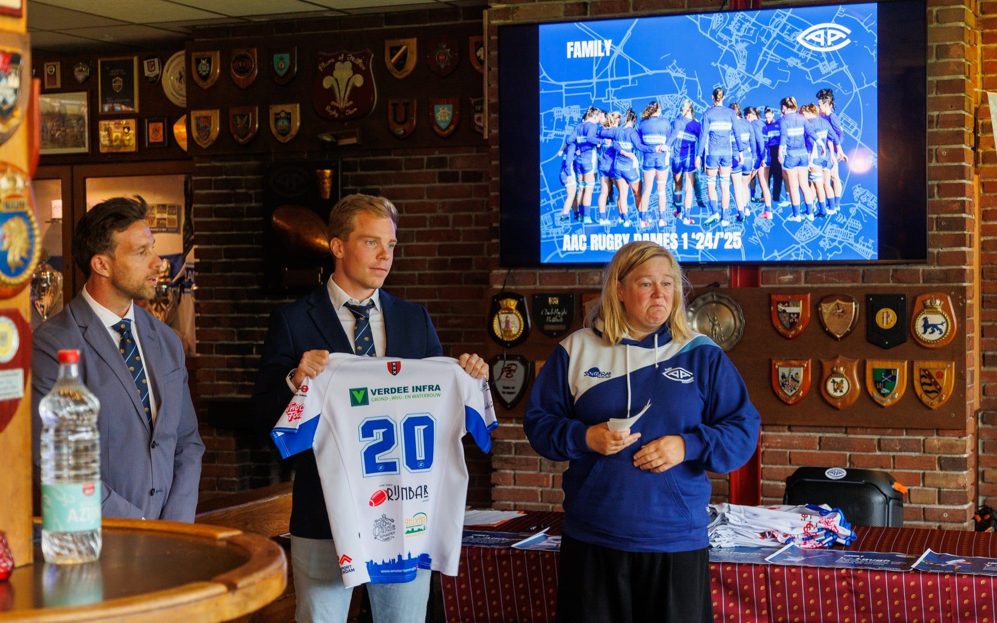 AAC-shirt met sponsorlogo in het clubhuis naast een scherm met het clubembleem en de selectie